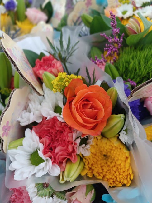 A vibrant floral bouquet featuring an orange rose, white daisies with green centers, pink carnations, yellow chrysanthemums, and purple lavender, arranged with green foliage and wrapped in light paste