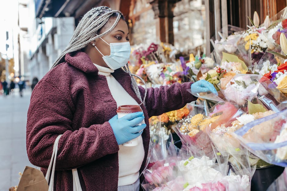 A woman with braided silver hair, wearing a face mask, blue gloves, and a burgundy fleece jacket, selecting a floral arrangement from an outdoor flower stall on Plumstead High Street. The display feat