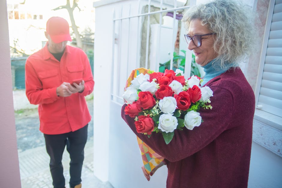A woman with curly gray hair, wearing glasses, a maroon sweater, and a light blue turtleneck, holding a vibrant bouquet of fresh red and white roses wrapped in colorful paper, standing near a white do