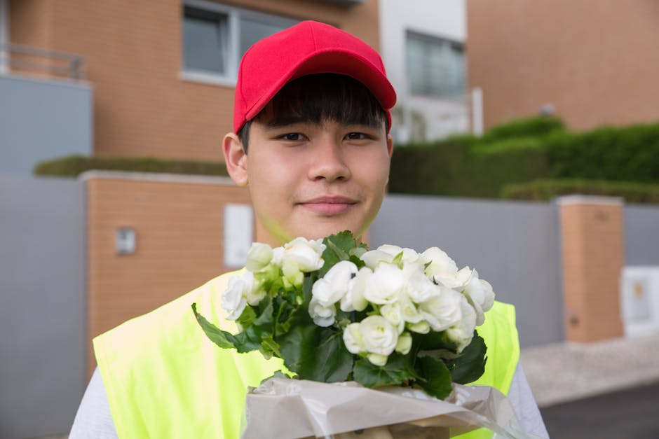 A young person wearing a red cap and a high-visibility vest is holding a bouquet of fresh white flowers with green foliage, wrapped in white paper. The bouquet features closely packed blooms with soft