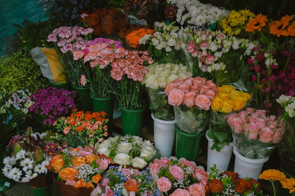 A colorful display of fresh flowers arranged in green buckets, white vases, and floral bouquets. The flowers include pink, peach, white, yellow, and purple blooms such as roses, carnations, daisies, a