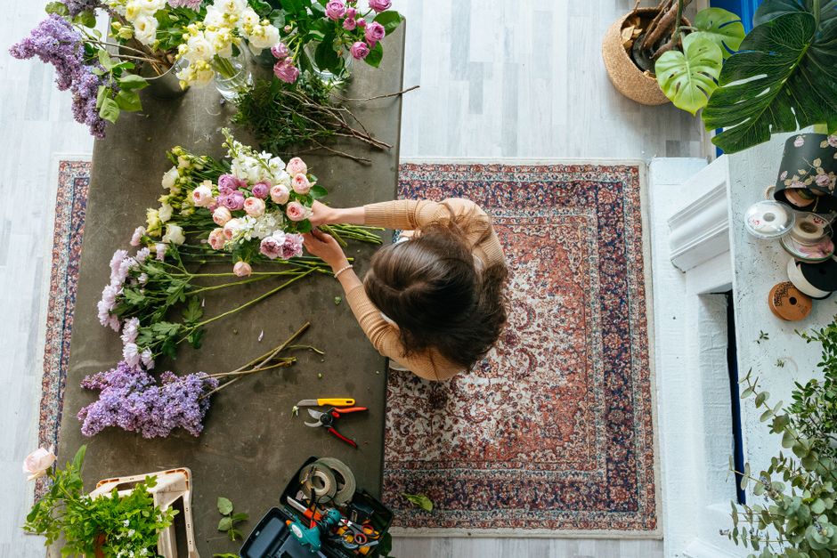 A woman arranging a floral bouquet on a rustic table in a bright, well-lit room. The bouquet includes soft pink roses, white lisianthus, purple lavender, and greenery. She is wrapping the flowers with